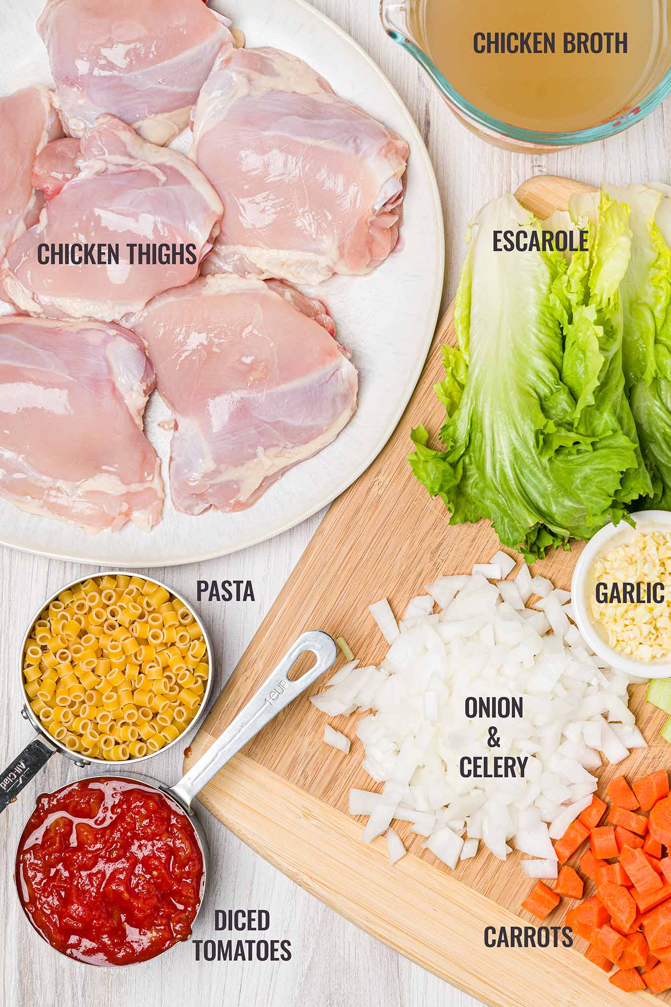 An overhead view of Italian chicken soup ingredients including skinless chicken thighs, broth, escarole, onion, celery, garlic, carrots, tomatoes, and pasta.