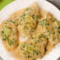Boneless chicken breasts on a white plate topped with a creamy sauce with chopped cilantro, and lime wedges.