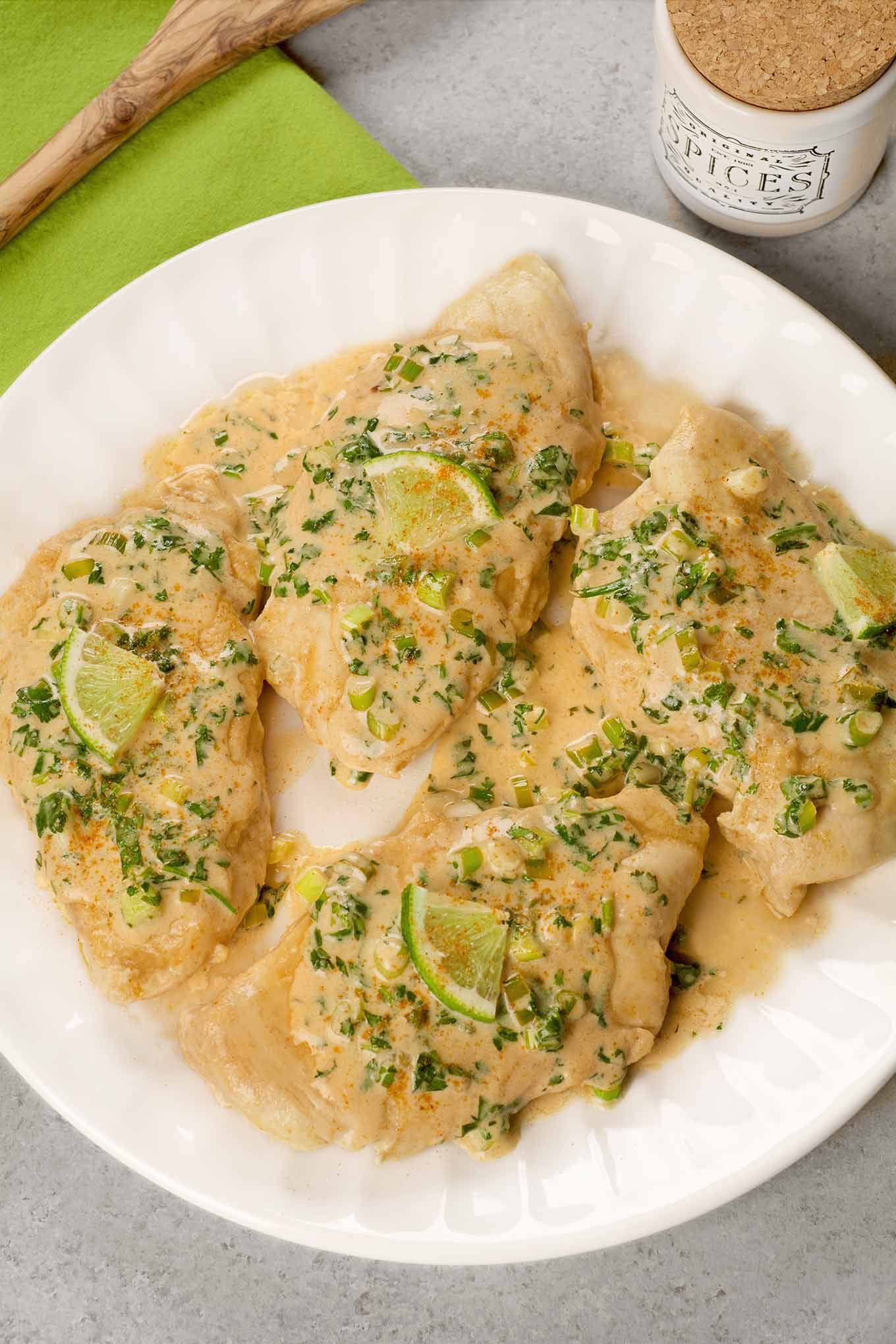 Boneless chicken breasts on a white plate topped with a creamy sauce with chopped cilantro, and lime wedges.