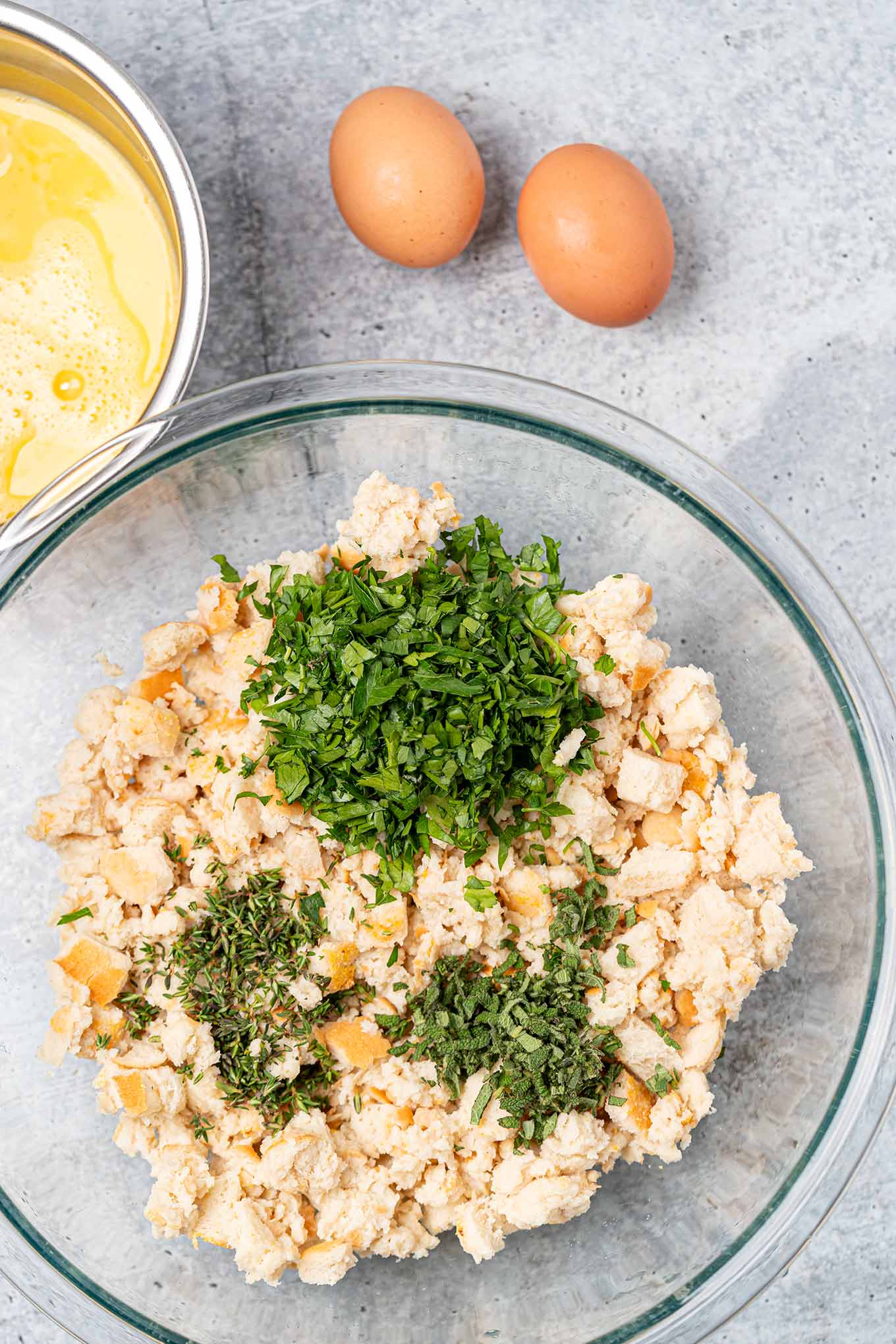Preparing bread stuffing, one mixing bowl with moistened bread cubes, fresh thyme, parsley, and sage, beaten eggs in a separate bowl to the side.