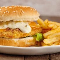 An oven-fried fish fillet on a seeded soft roll with melted cheddar cheese, tartar sauce, and lettuce, and fries on the plate to the right.