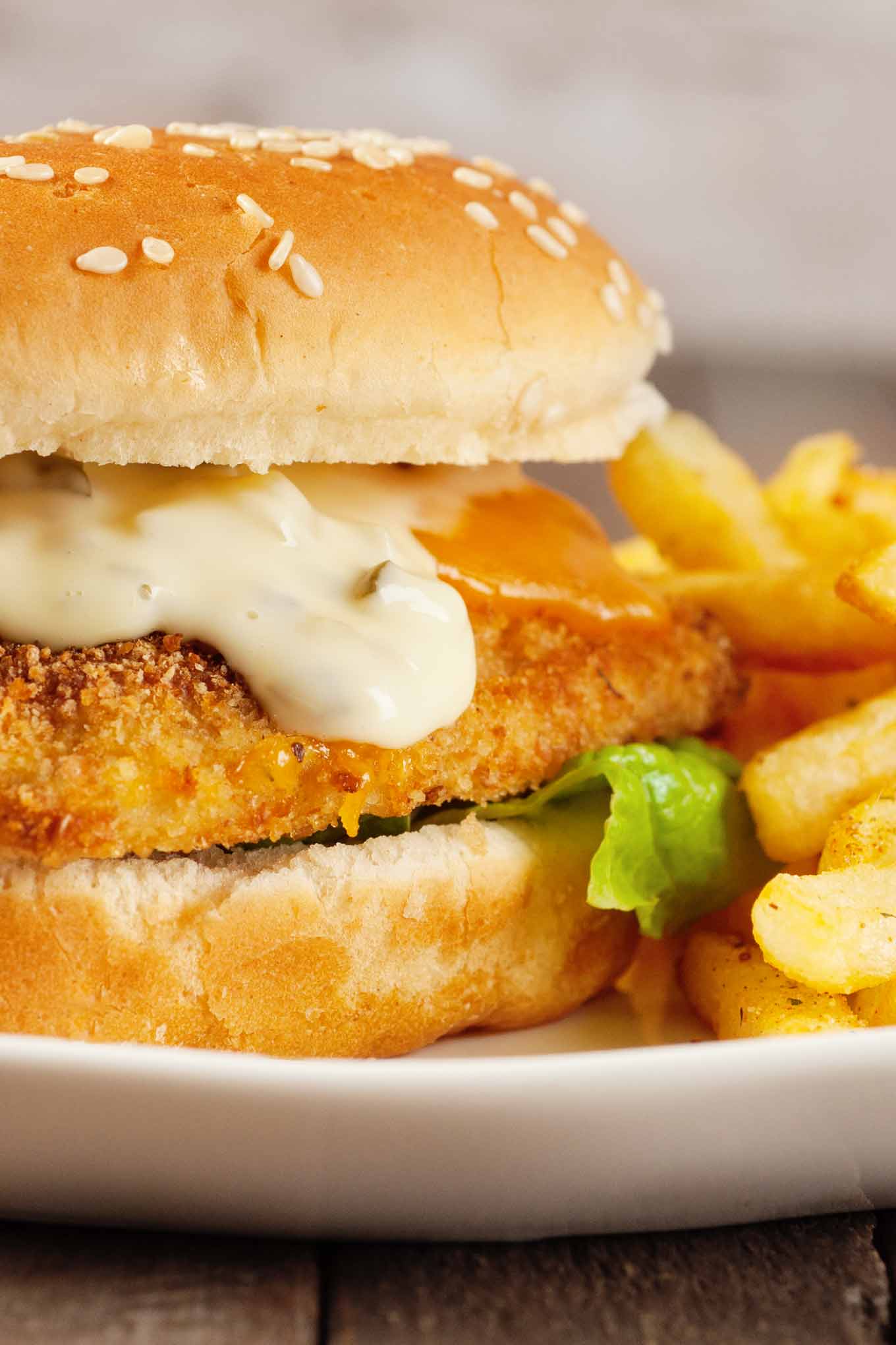 A close-up view of an oven-fried fish fillet on a seeded roll with melted cheese, tartar sauce, and lettuce, and fries on the plate to the right.