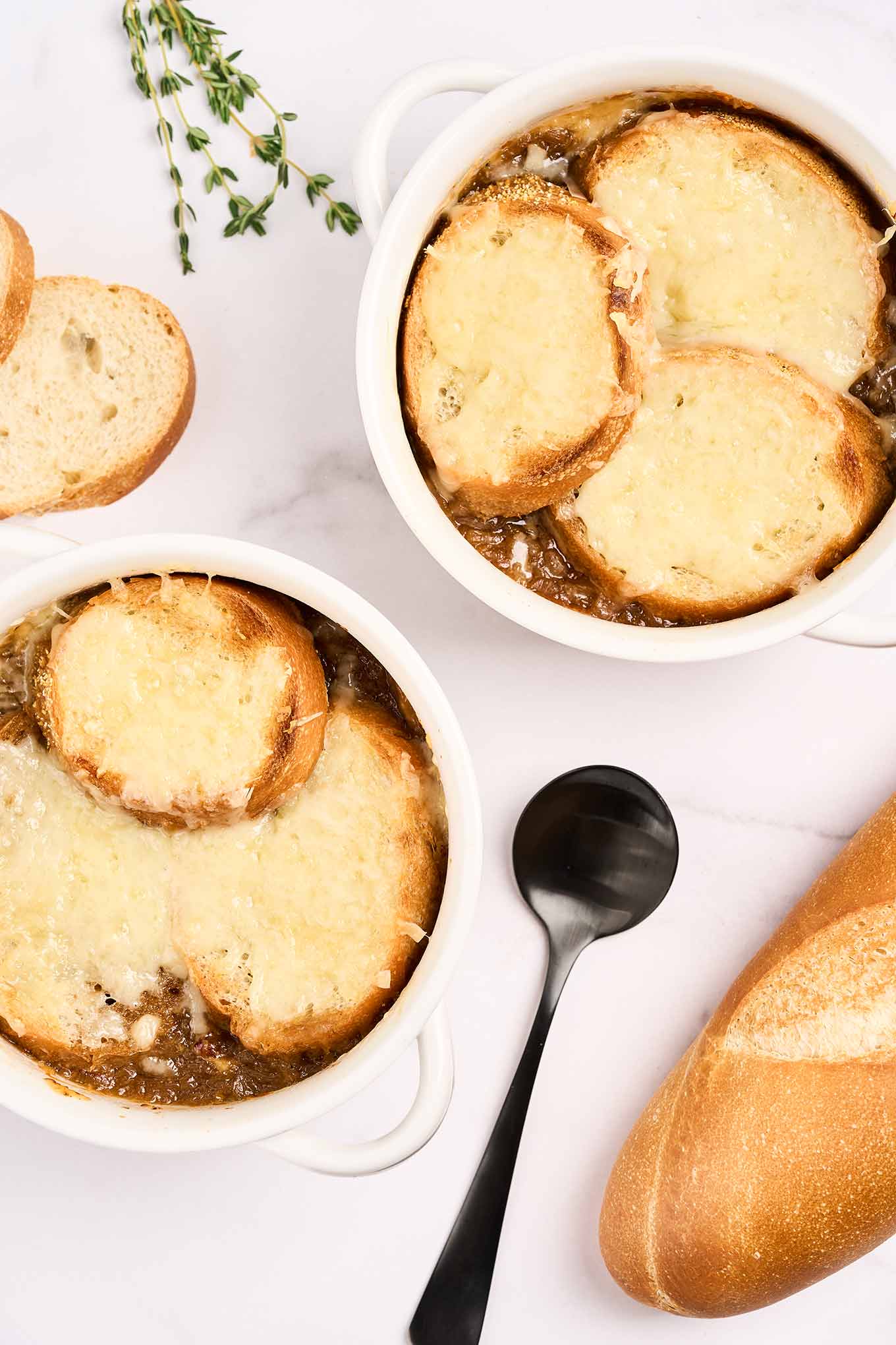 Two white soup crocks filled with French onion soup topped with baguette and melted cheese surrounded by fresh thyme, unsliced baguette, and a spoon.