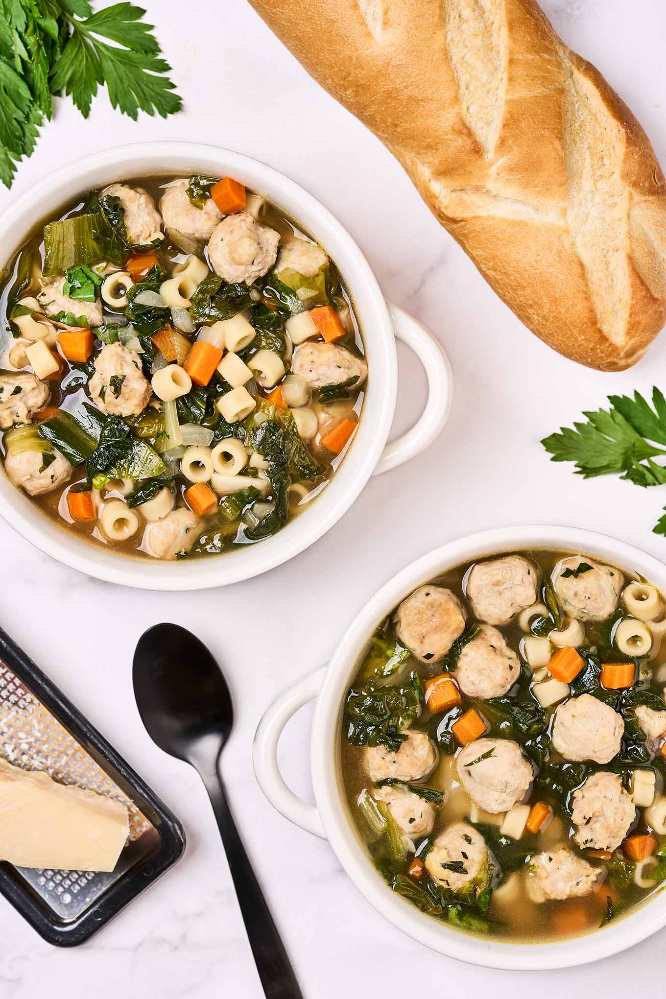Two white soup crocks filled with chicken soup with ditalini pasta, escarole, and mini turkey meatballs surrounded by sprigs of fresh parsley, a loaf of French bread, a black spoon, a cheese grater, and a block of Parmesan cheese.