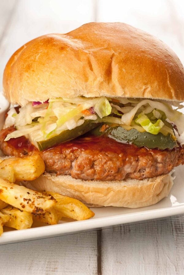 A close view of a grilled pork patty topped with bourbon bbq sauce, pickle slices, and coleslaw on a soft hamburger roll with fries on the side.