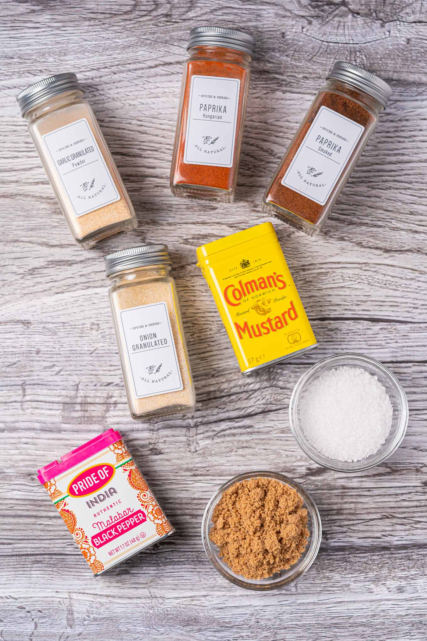 An overhead view of the ingredients for BBQ spice rub; sweet and smoked paprika, brown sugar, salt, granulated garlic and onion, pepper, and dry mustard.