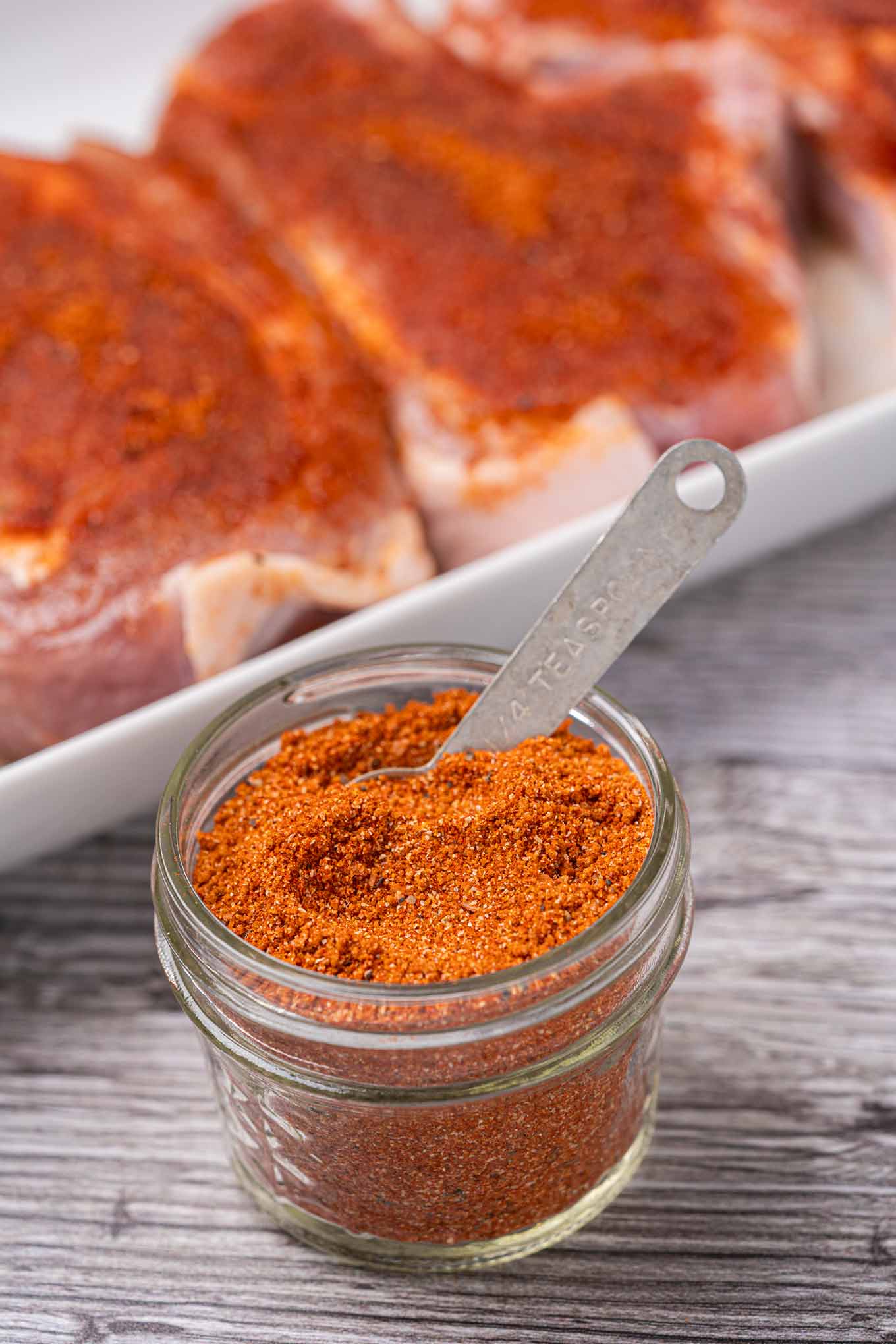 A glass jar of BBQ spice rub with a measuring spoon in it with rub-coated pork chops in the background.