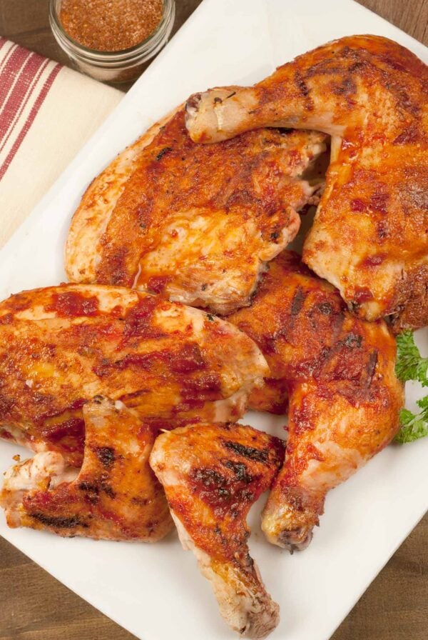 A white serving platter with bbq chicken parts, leg quarters, breasts, and wings. A sprig of parsley on the side and a jar of bbq spice rub in the background.