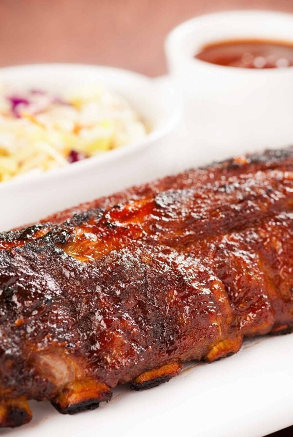 A close side view of a whole rack of baby back ribs, oven baked, glazed with homemade bourbon bbq sauce, coleslaw and more bbq sauce in the background.