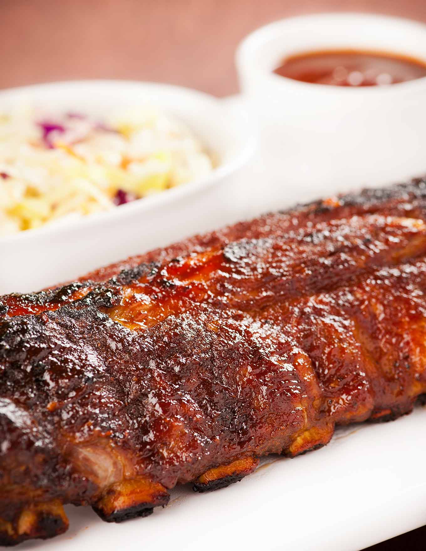 A close side view of a whole rack of baby back ribs, oven baked, glazed with homemade bourbon bbq sauce, coleslaw and more bbq sauce in the background.