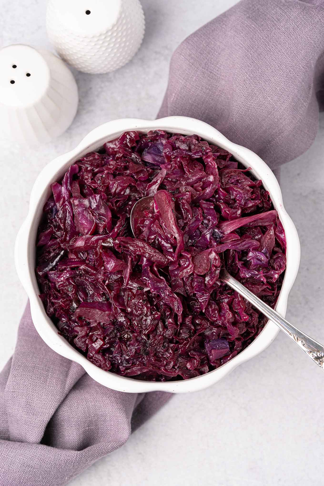 An overhead view of a serving bowl of braised red cabbage with a spoon in it, on top of a linen napkin with white ceramic salt and pepper shakers behind it and to the left.