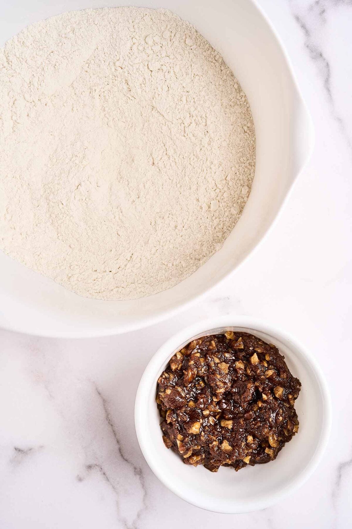 A mixing bowl containing the dry ingredients for making cinnamon walnut bread with a bowl of chopped walnuts mixed with melted butter and cinnamon beside it.