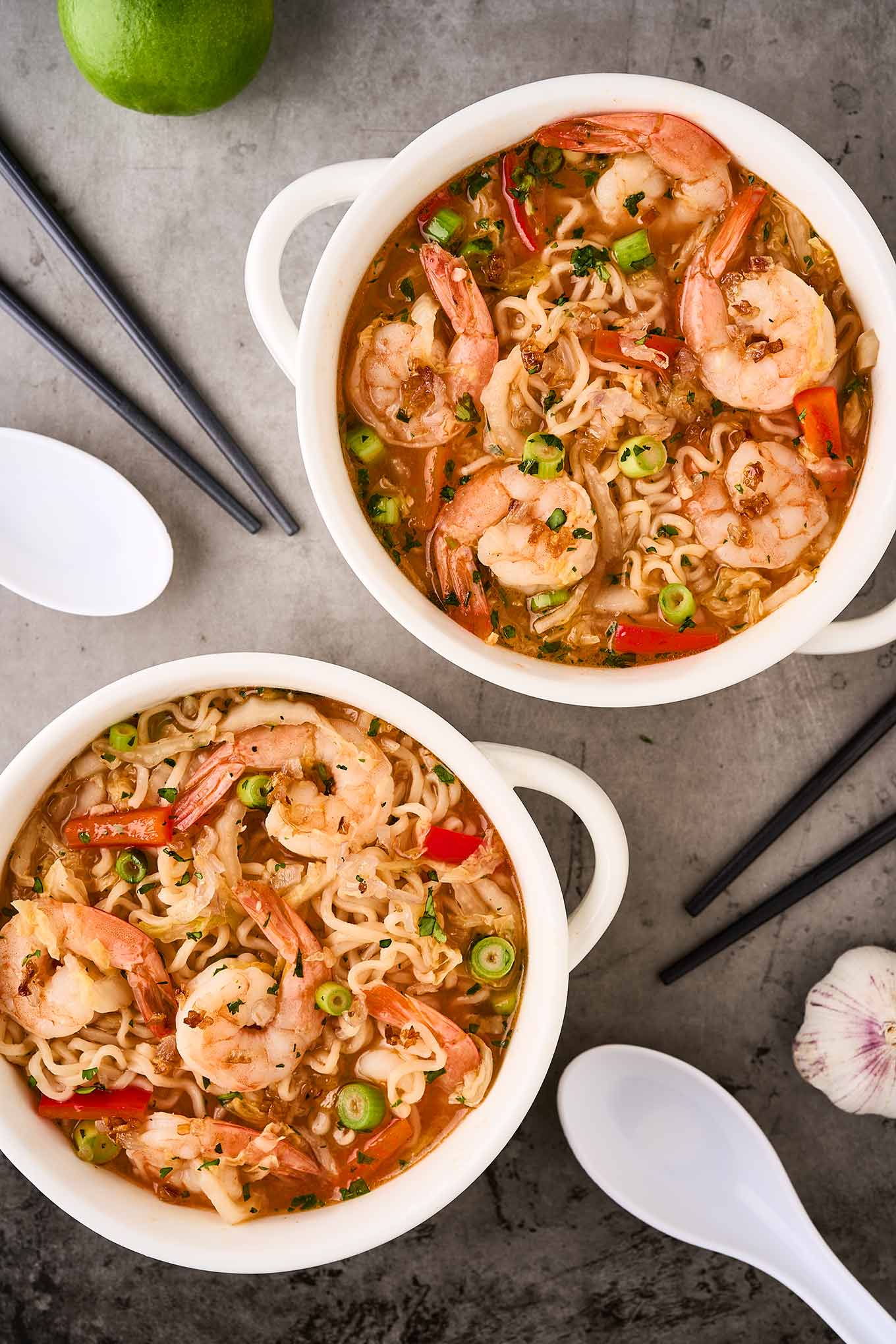 An overhead view of two bowls of shrimp ramen noodle soup surrounded by chopsticks, soup spoons, a whole lime, and a head of garlic.