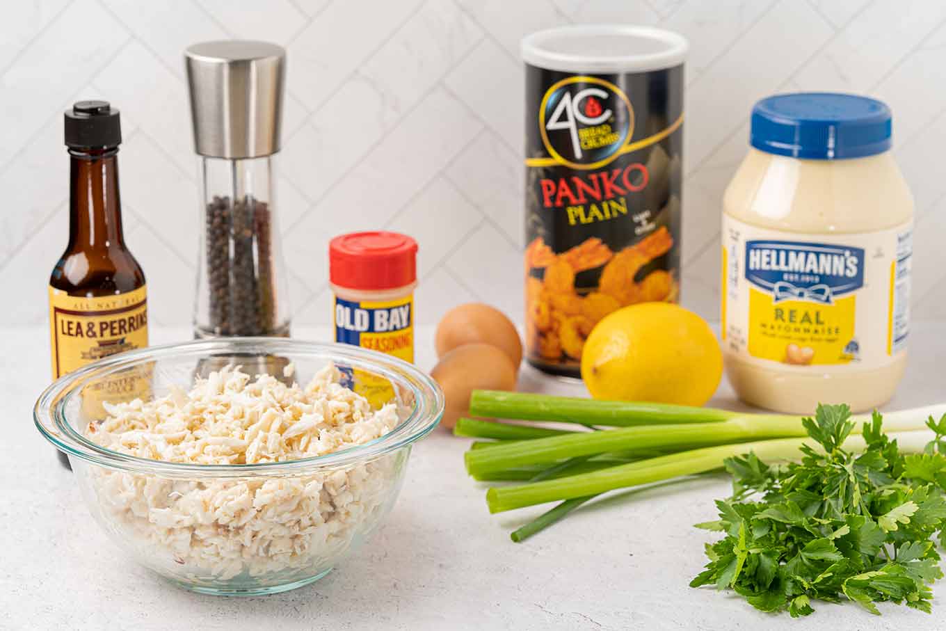 The ingredients for baked crab cakes arranged on a counter; crabmeat, eggs, mayonnaise, panko crumbs, Old Bay Seasoning, lemon, scallions, parsley, black pepper, and Worcestershire sauce.