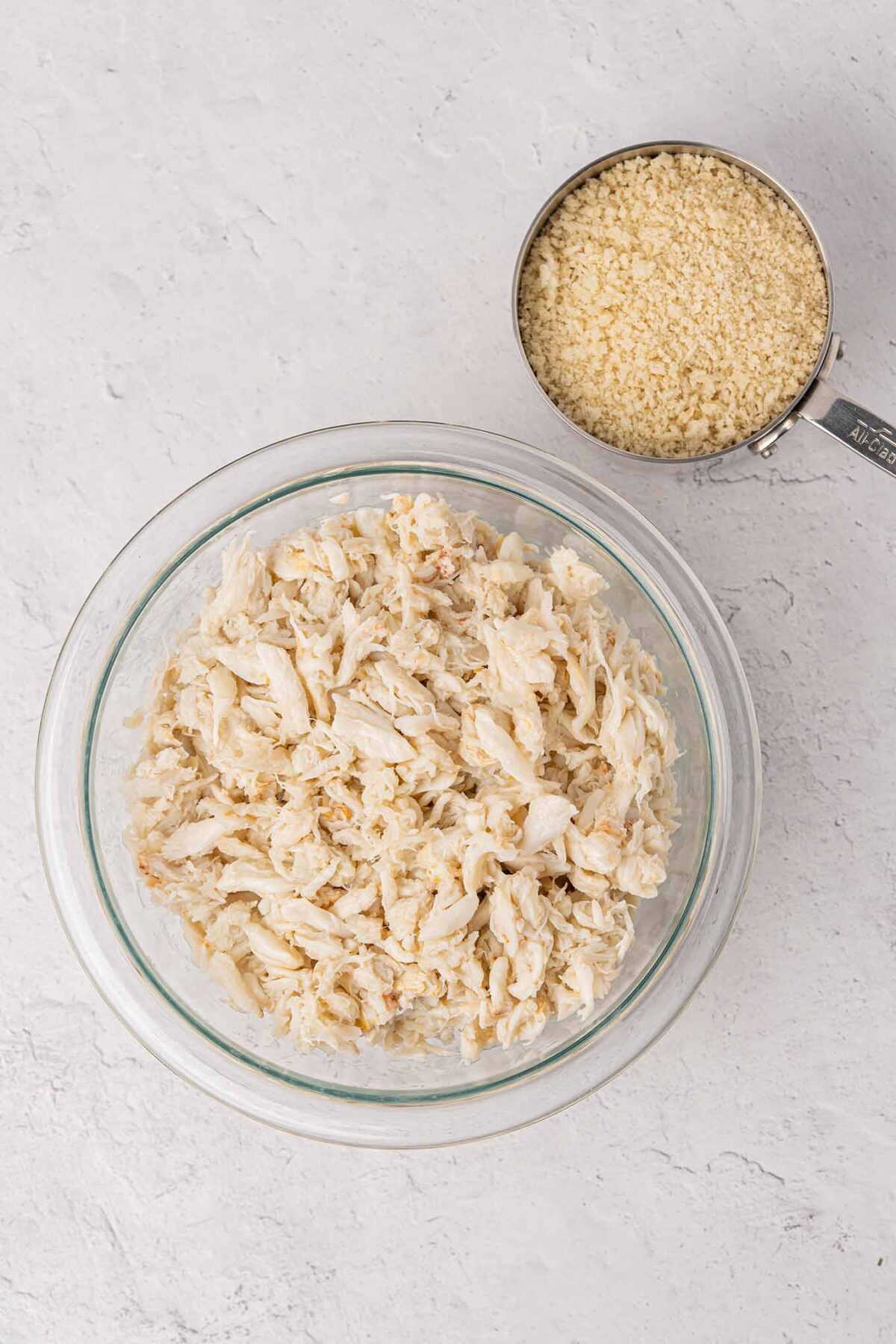 Lump crabmeat in a glass bowl next to a measuring cup filled with panko crumbs.