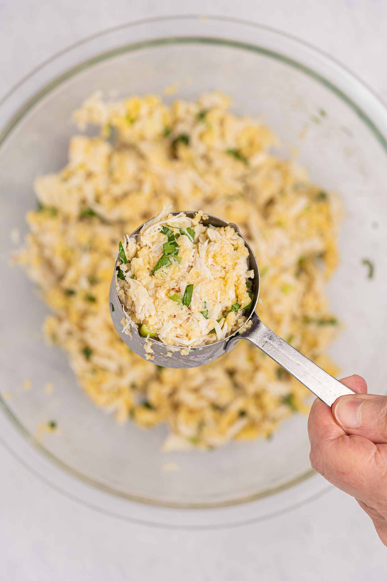 An overhead view of crab cake mixture packed into a metal measuring cup being held over a mixing bowl with more.