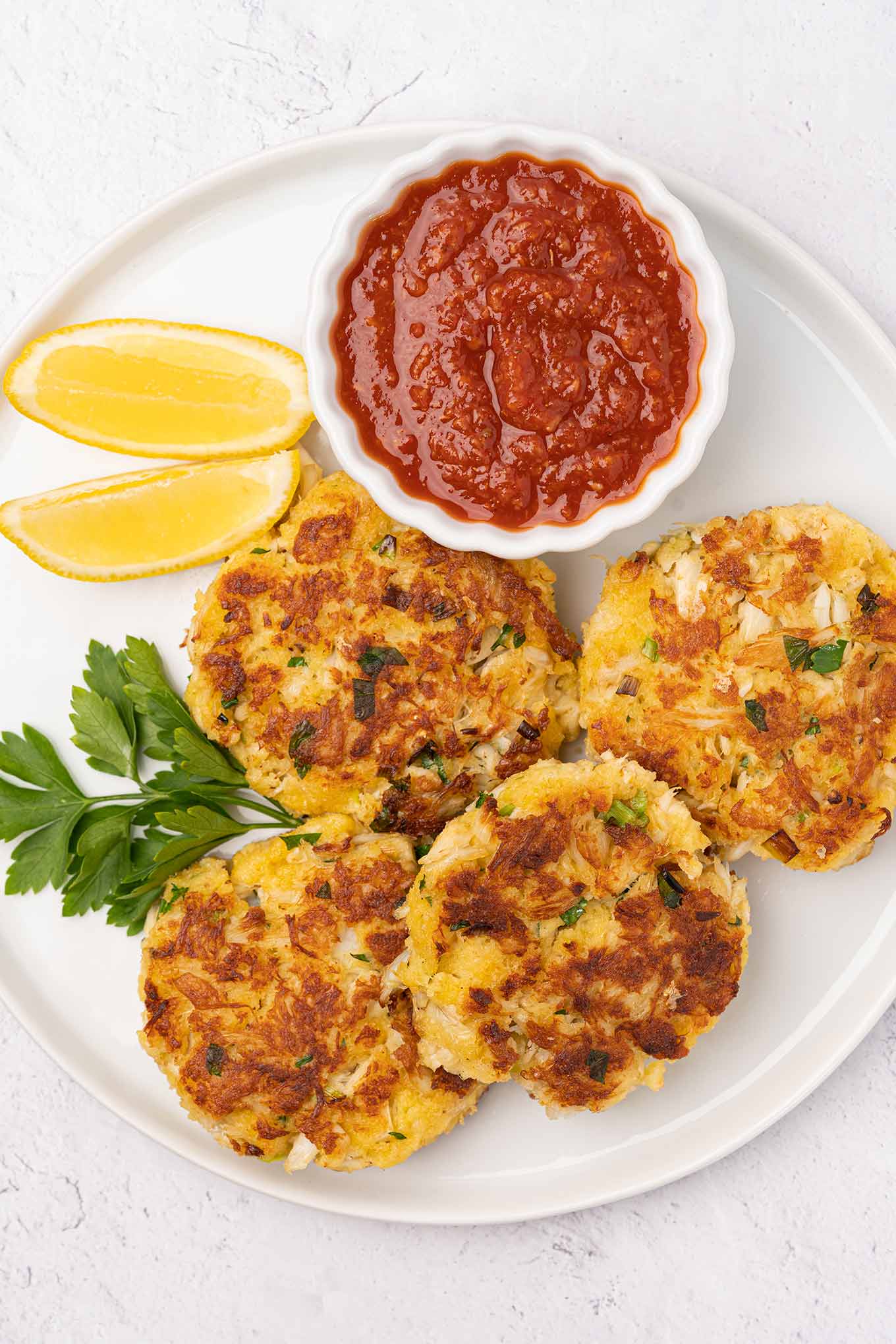 An overhead view of four crispy crab cakes on a round plate with lemon wedges and a bowl of cocktail sauce.