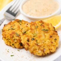 A close-up of two crisp, golden brown, baked crab cakes on a plate, a fork and yellow napkin to the side, lemon and remoulade sauce in the background.