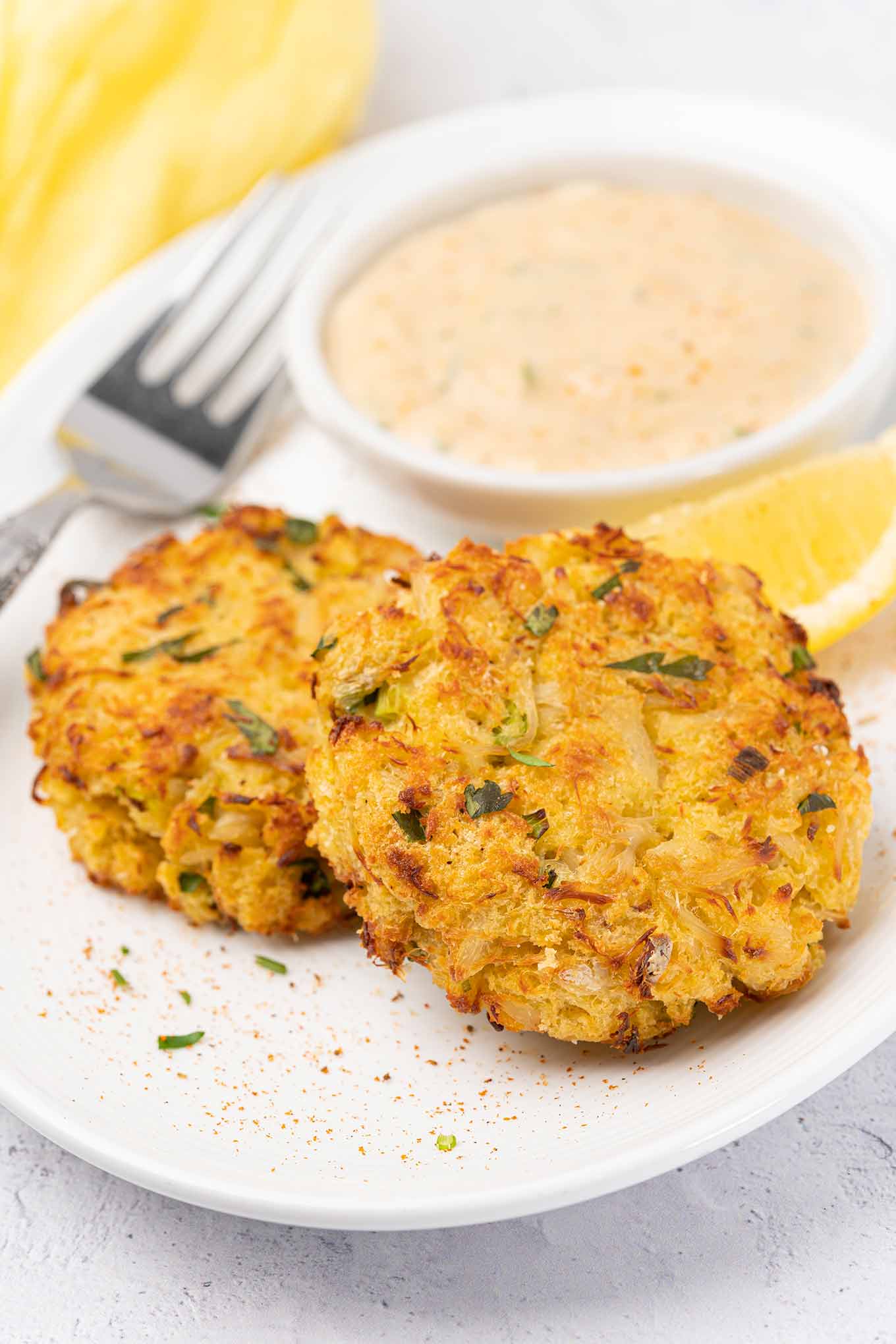 A close-up of two crisp, golden brown, baked crab cakes on a plate, a fork and yellow napkin to the side, lemon and remoulade sauce in the background.