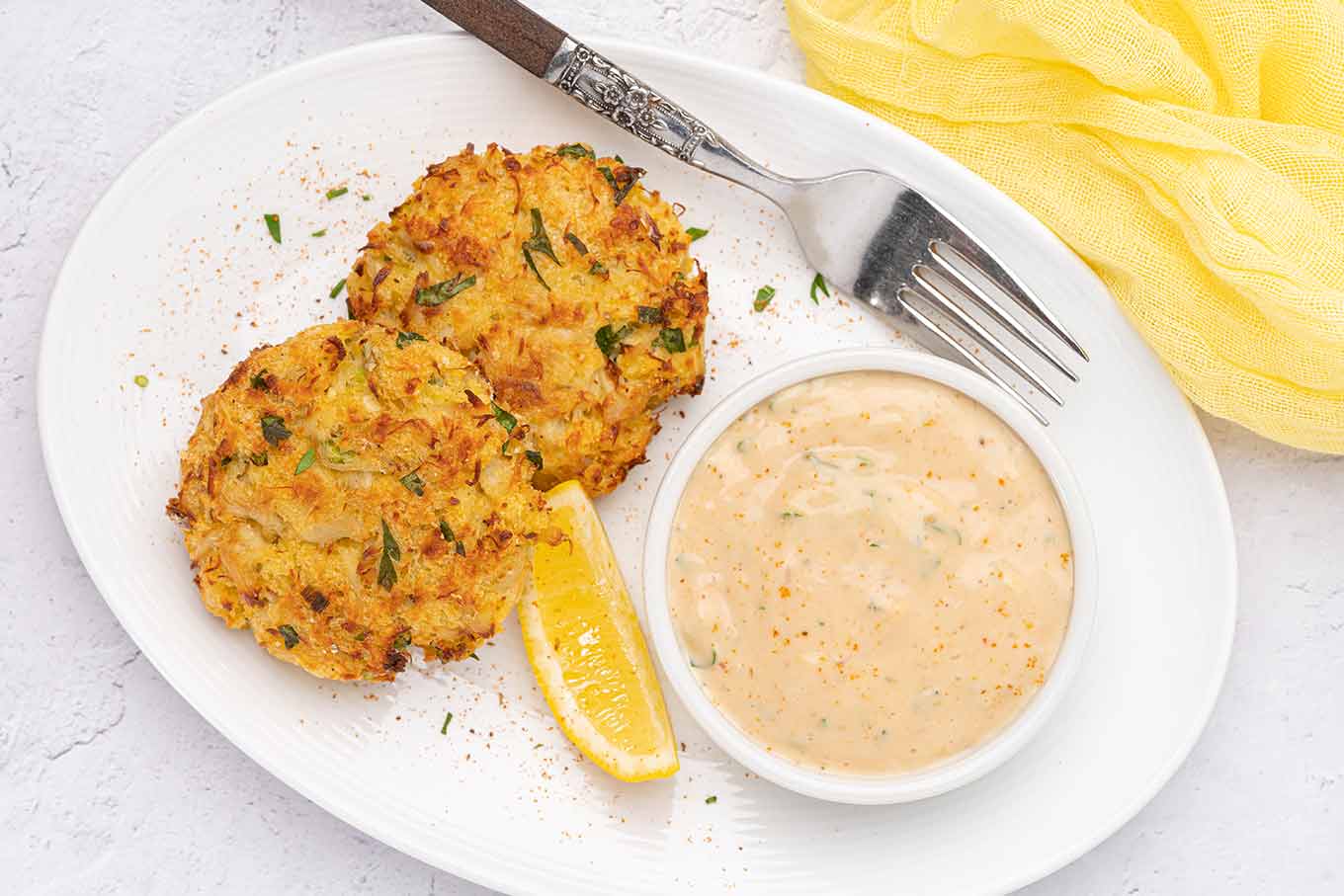 An overhead view of two baked crab cakes on an oval serving plate with a lemon wedge, fork, and bowl of remoulade for dipping.