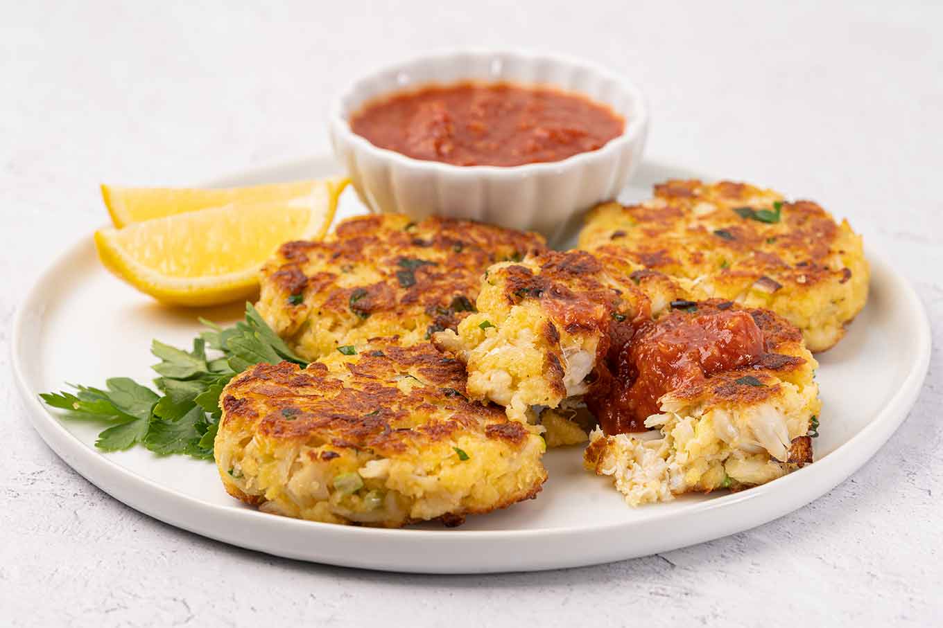 A close view of a serving plate with four crab cakes, one cut to show texture, surrounded by lemon wedges, parsley, and a small bowl of cocktail sauce.