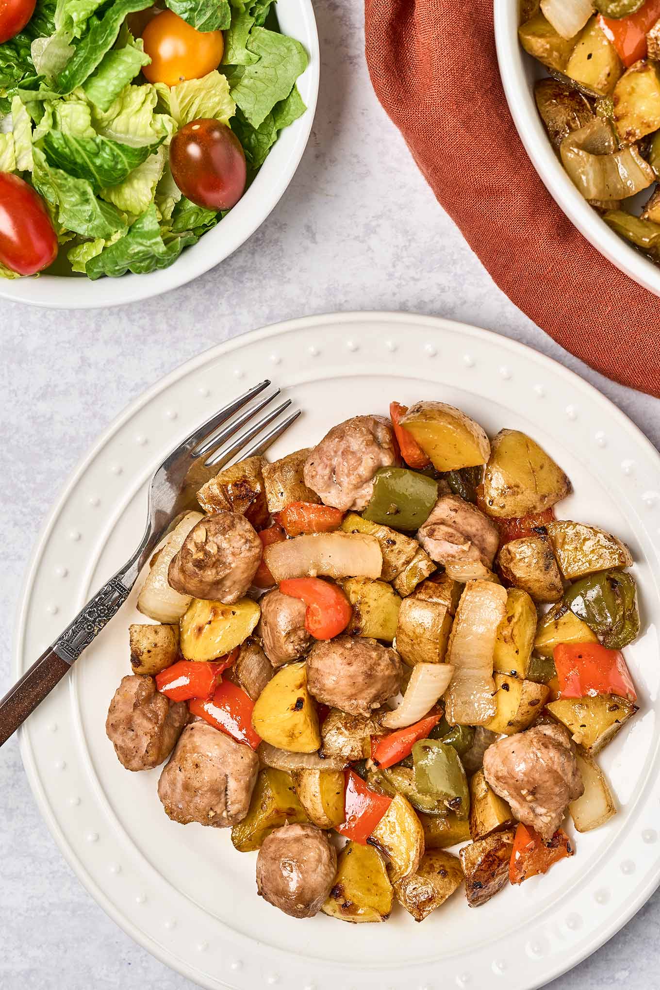A round white dinner plate with with balsamic glazed baked Italian sausage, bell peppers, and onions, a dinner fork to the side and a serving bowl with more of the main dish and a bowl of salad with grape tomatoes in the background.