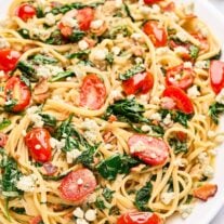 A serving platter of blue cheese pasta with caramelized onions, bacon, fresh spinach, and tomatoes, a block of blue cheese, fresh basil, and halved grape tomatoes in the background.