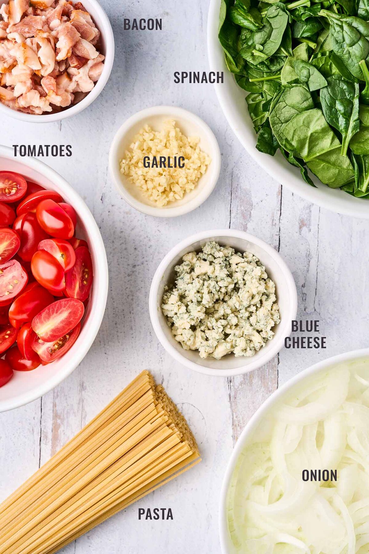 A labeled, overhead photo of the ingredients for blue cheese pasta; bacon, fresh spinach, grape tomatoes, garlic, crumbled blue cheese, onion, and linguine.