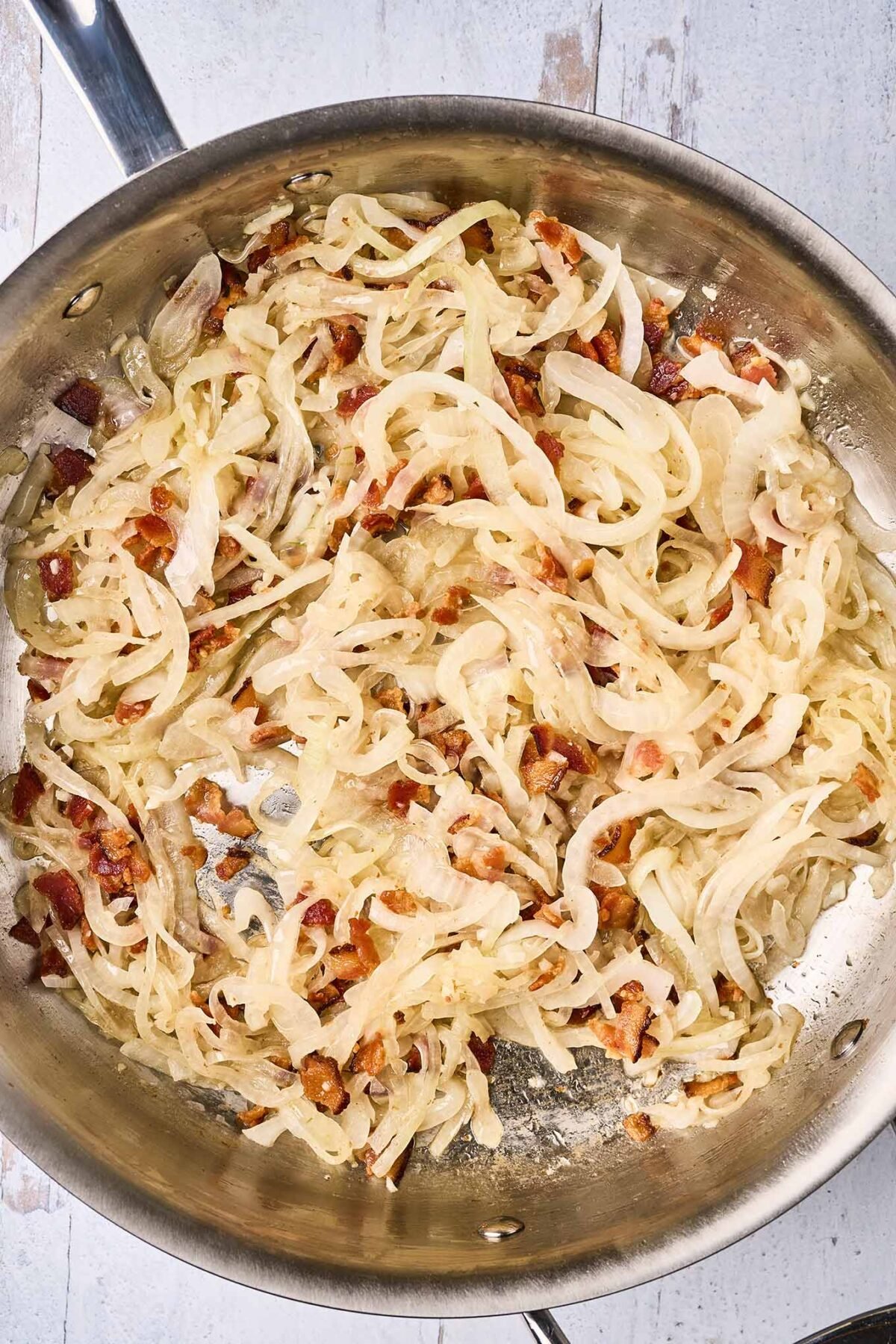 A sauté pan with bits of crispy, thick-cut bacon and sliced sweet onion beginning to caramelize.
