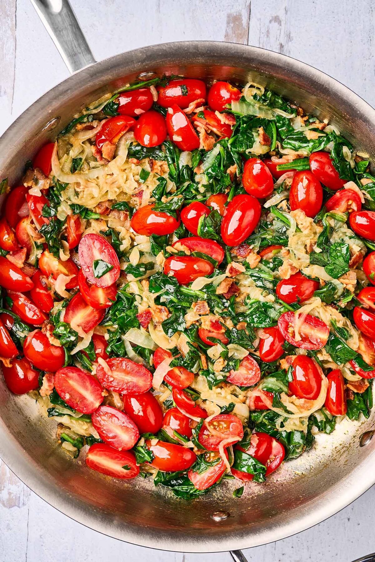 A sauté pan filled with caramelized onions, bacon, sautéed grape tomatoes, and sautéed spinach, ready to be combined with cooked linguine pasta.