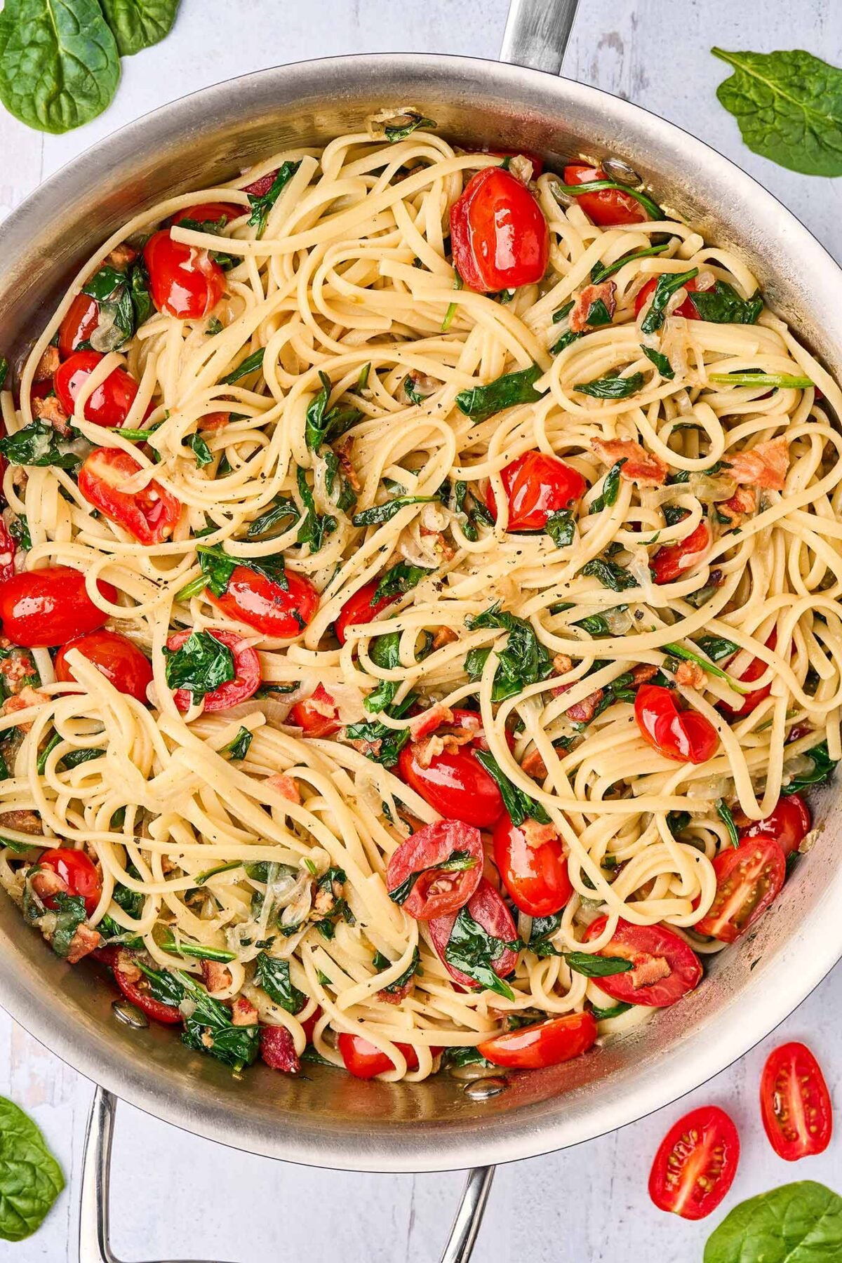 A sauté pan filled with linguine tossed with caramelized onions, bacon, fresh spinach, and halved grape tomatoes, ready for the addition of crumbled blue cheese.