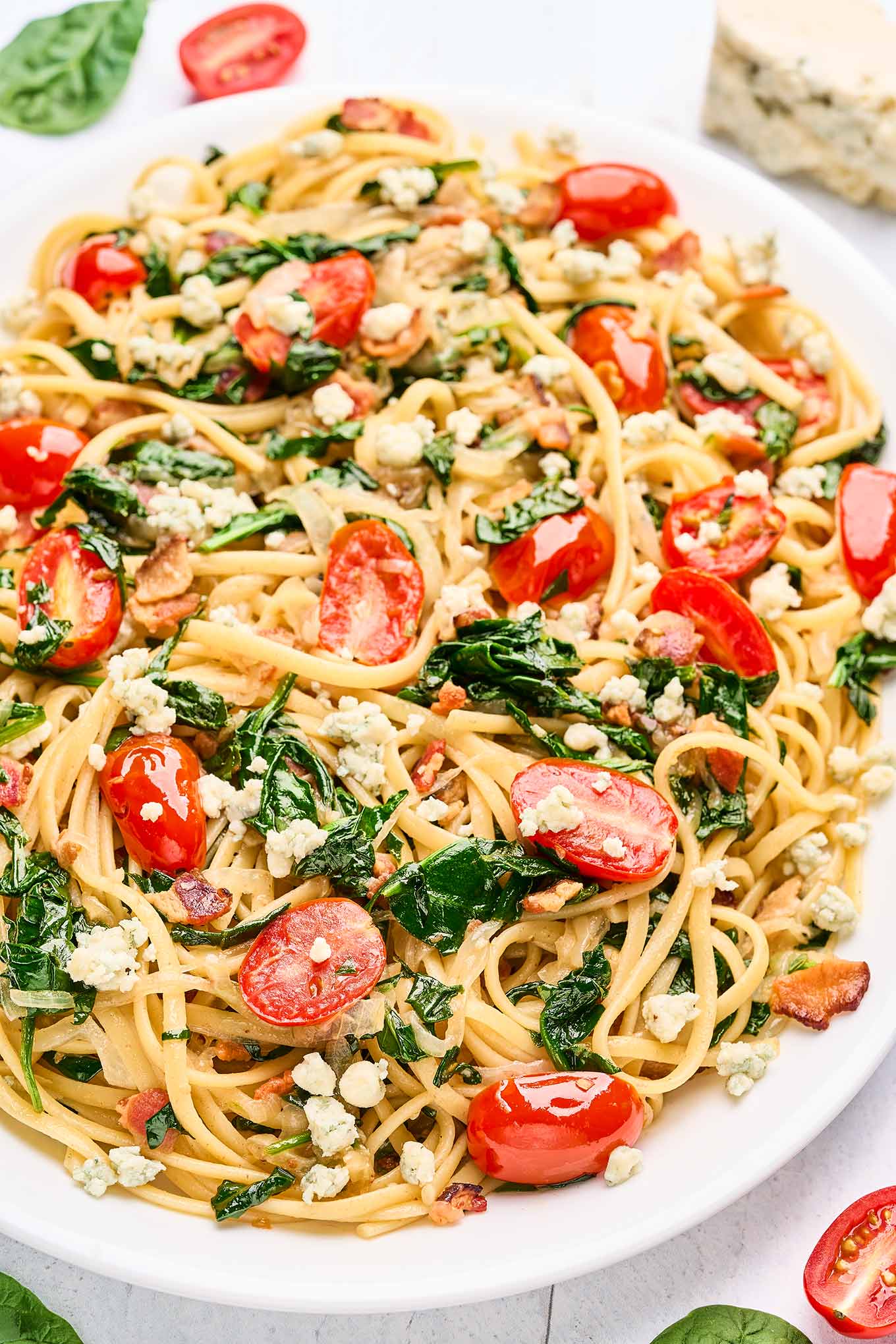 A serving platter of blue cheese pasta with caramelized onions, bacon, fresh spinach, and tomatoes, a block of blue cheese, fresh basil, and halved grape tomatoes in the background.