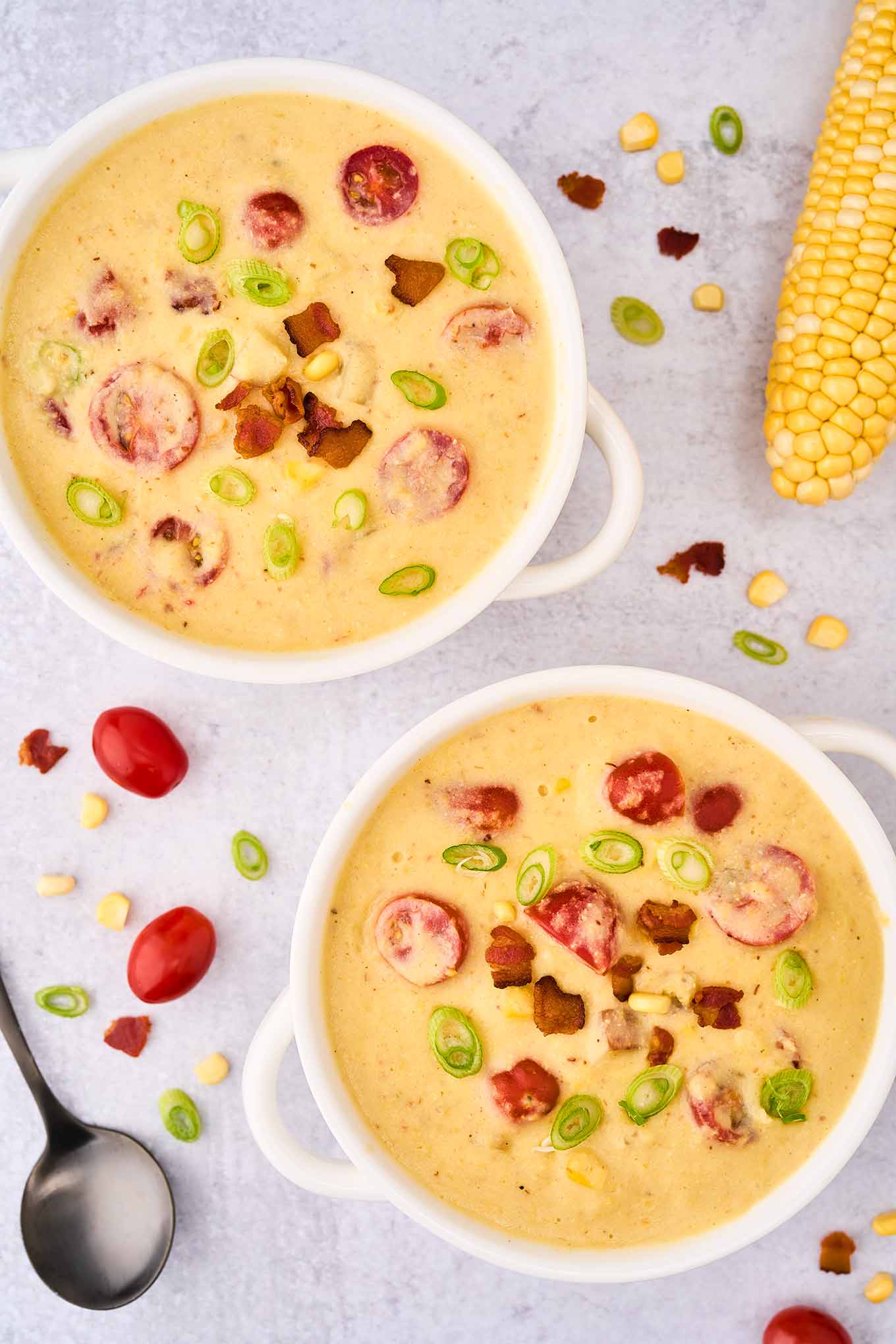 An overhead view of two white soup crocks of corn chowder garnished with bacon, tomatoes, corn, and scallions, a whole ear of corn, a spoon, and a scattering ingredients in the background.