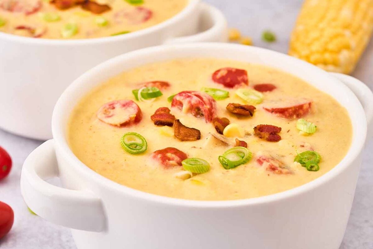A close up of a white soup crock of corn chowder garnished with crispy bacon, sliced scallions, corn kernels, and halved grape tomatoes.