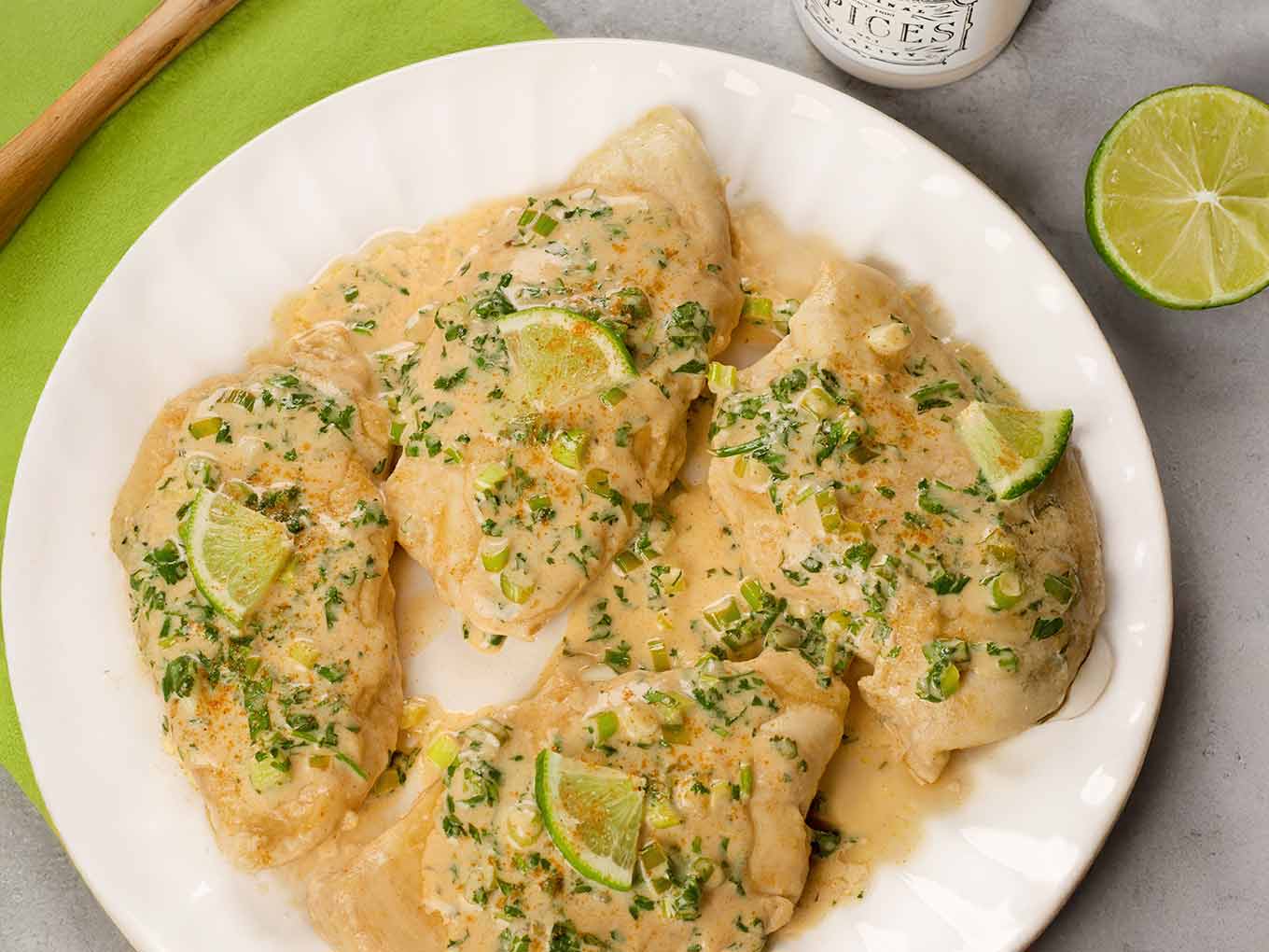 Four pan-seared, boneless chicken breasts topped with creamy cilantro lime sauce, scallions, and lime slices, surrounded by a green napkin, spice jar, and lime half.