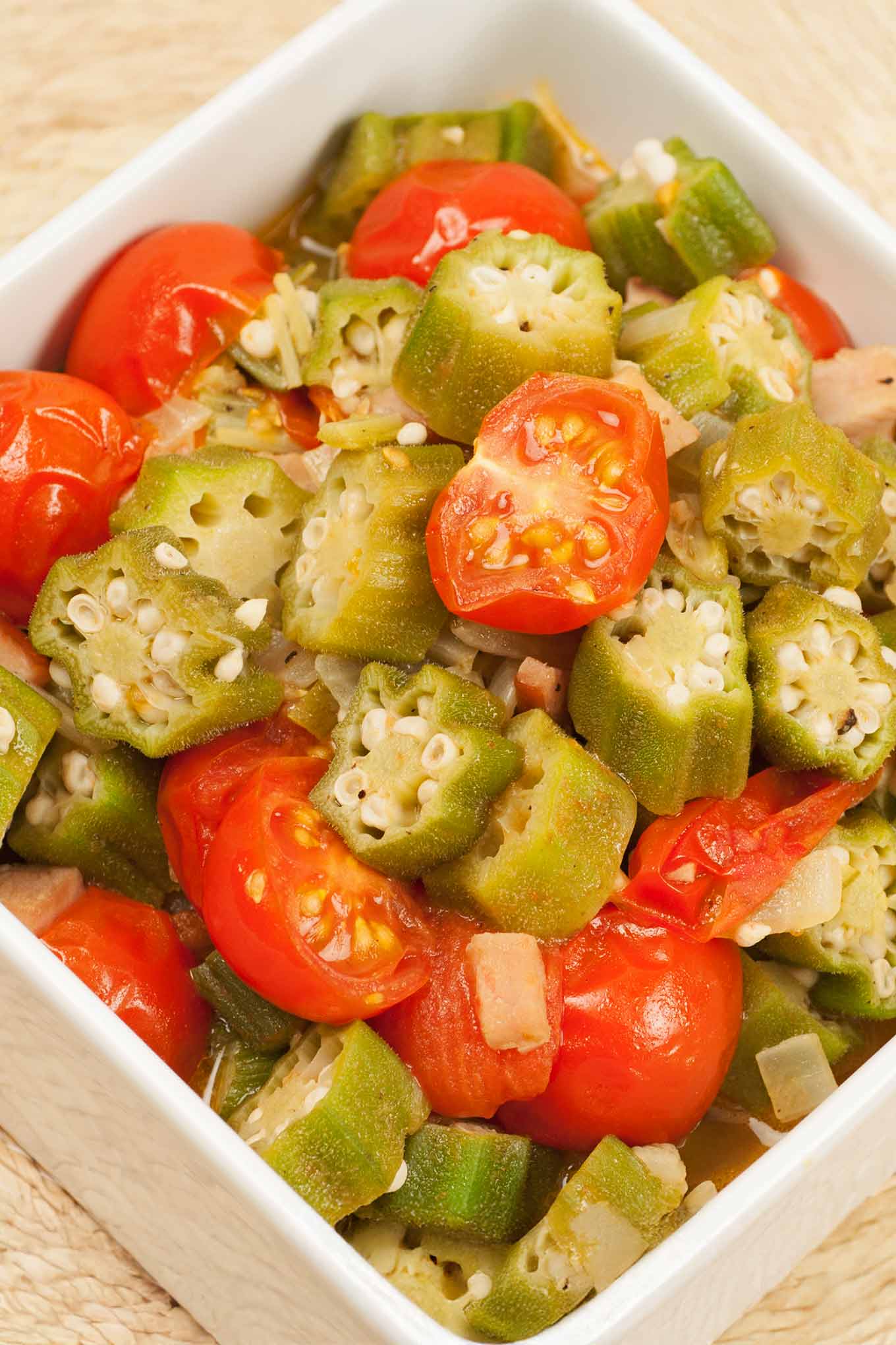 A square, white serving bowl with chunks of okra and halved cherry tomatoes sautéed with onion and garlic in olive and butter.