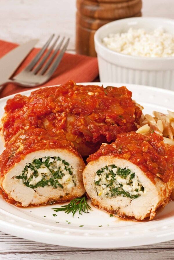 A dinner plate with boneless chicken breasts, rolled and stuffed with spinach and feta cheese, and topped with Greek-style tomato sauce, utensils, a small bowl of feta cheese, and a pepper mill in the background.