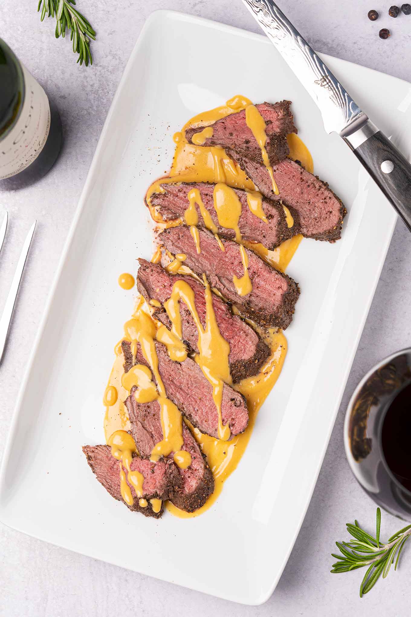 An overhead view of a white serving platter with slices of spice-crusted, roast venison tenderloin, drizzled with gjetost cheese sauce, a carving knife on the edge of the plate, and sprigs of fresh rosemary, peppercorns, and a glass of red wine off to the side.
