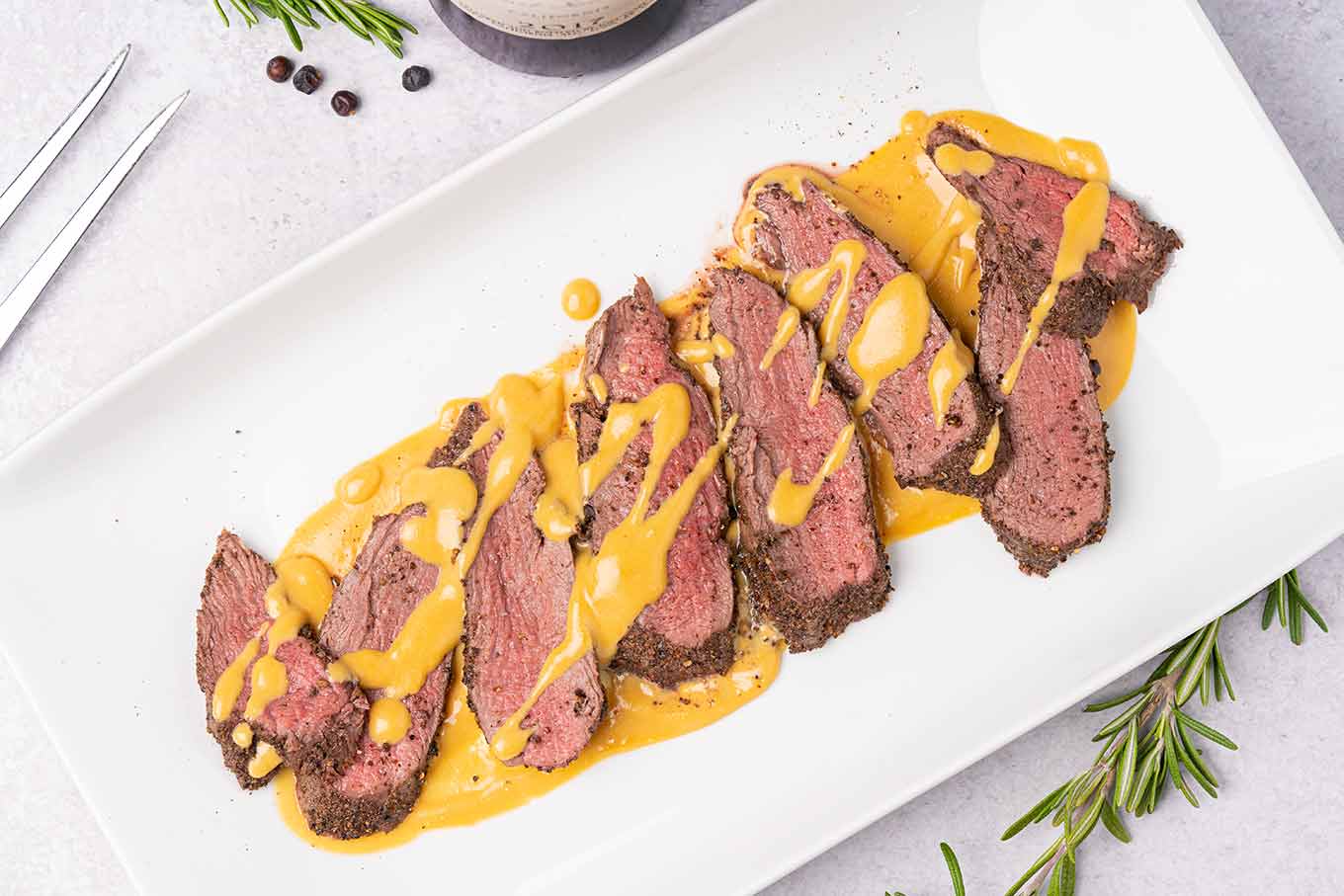 A white oblong platter arranged with slices of medium-rare roasted venison tenderloin drizzled with gjetost cheese sauce.