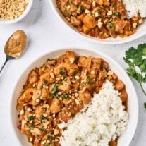 An overhead view of two bowls of West African chicken peanut stew with white rice, topped with chopped peanuts and cilantro; a bowl of extra peanuts, a spoon of peanut butter, and a sprig of cilantro to the side.
