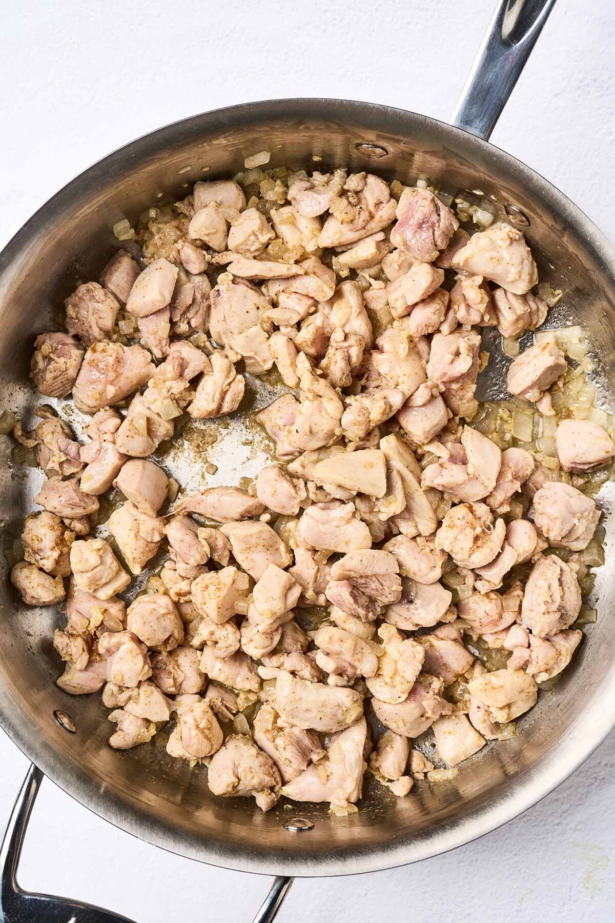 Bite sized chunks of boneless chicken thighs cooking in a sauté pan with chopped onion, garlic, and ginger.