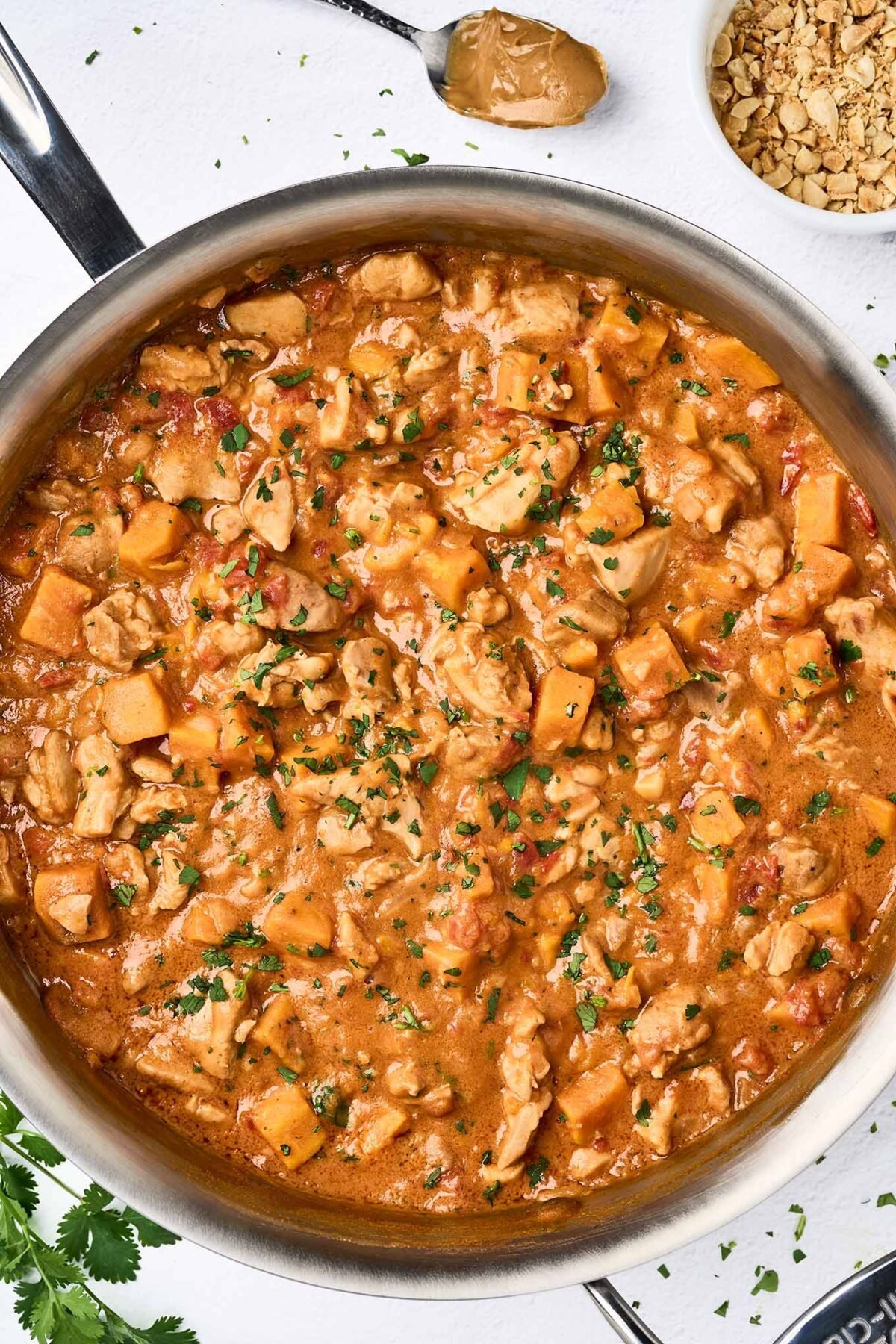 A sauté pan filled with African peanut stew with chunks of chicken thighs, sweet potato in a thick, savory, peanut butter and tomato based sauce.