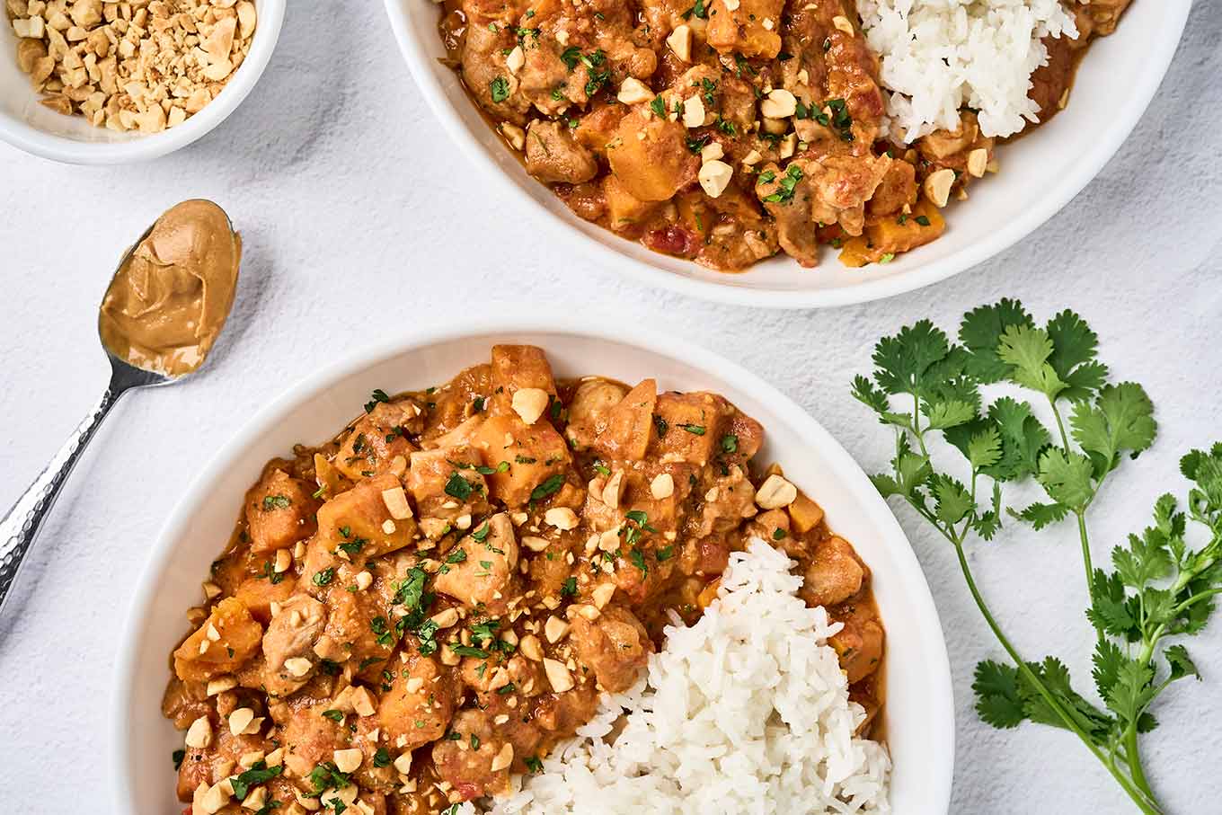 Two servings of West African chicken peanut stew and steamed white rice in bowls, garnished with chopped peanuts and cilantro, a dish with extra peanuts to the side.