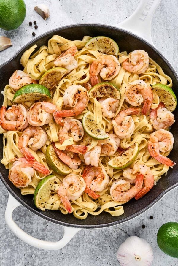 A cast iron pan filled with fettuccine tossed with sautéed shrimp, pan-seared lime wedges, and a light and garlicky white wine sauce.