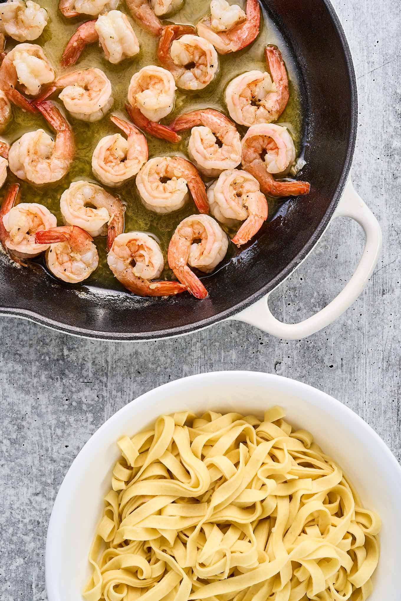 A cast iron pan with sautéed shrimp in butter sauce next to a bowl of fresh fettuccine.