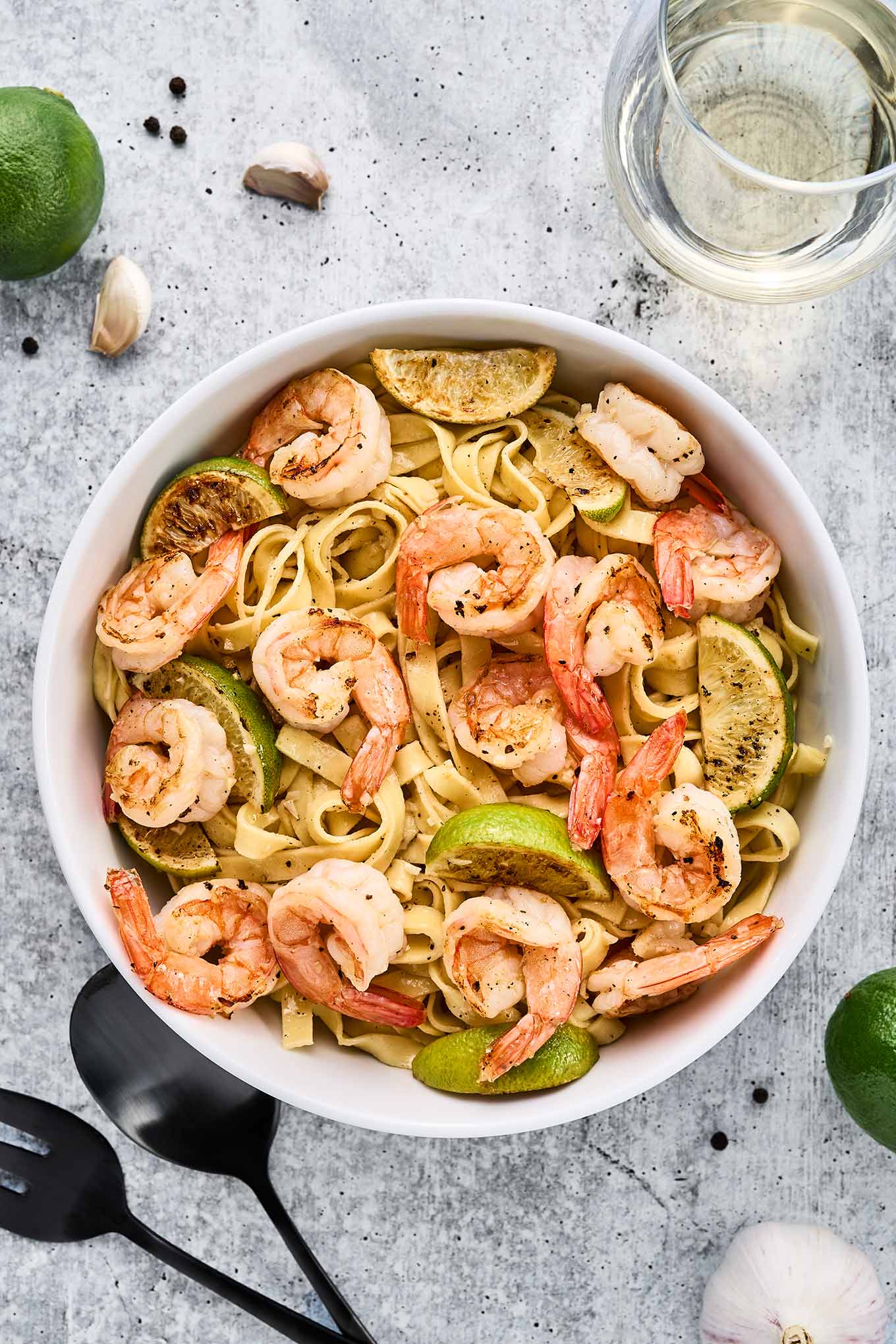 Garlic lime shrimp pasta garnished with lime wedges in a white bowl on a light stone surface, surrounded by garlic cloves, peppercorns, limes, and white wine.