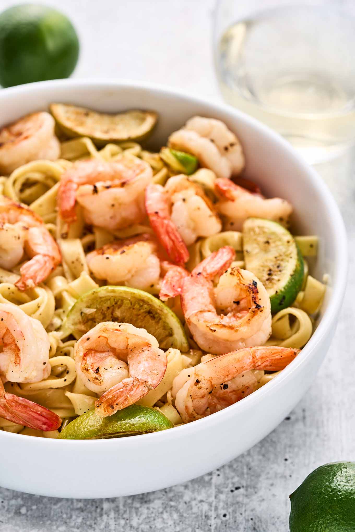 Close view of garlic lime shrimp tossed with fettuccine, showcasing the plump shrimp, pan-seared lime wedges, and tender pasta in a white bowl.