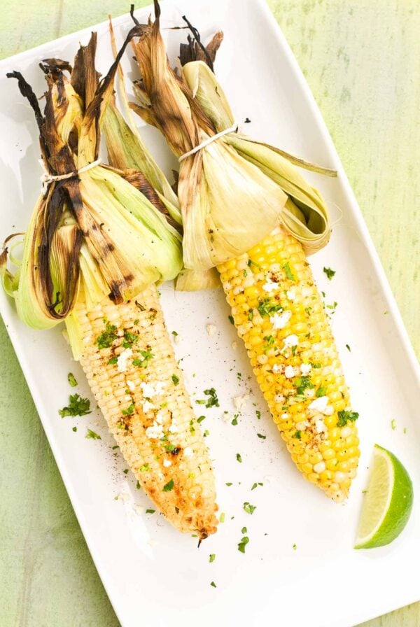 A serving platter with two ears of grilled corn, husks tied back, one white corn, one yellow, both topped with elote seasoning, chopped cilantro, and crumbled Cotija cheese.