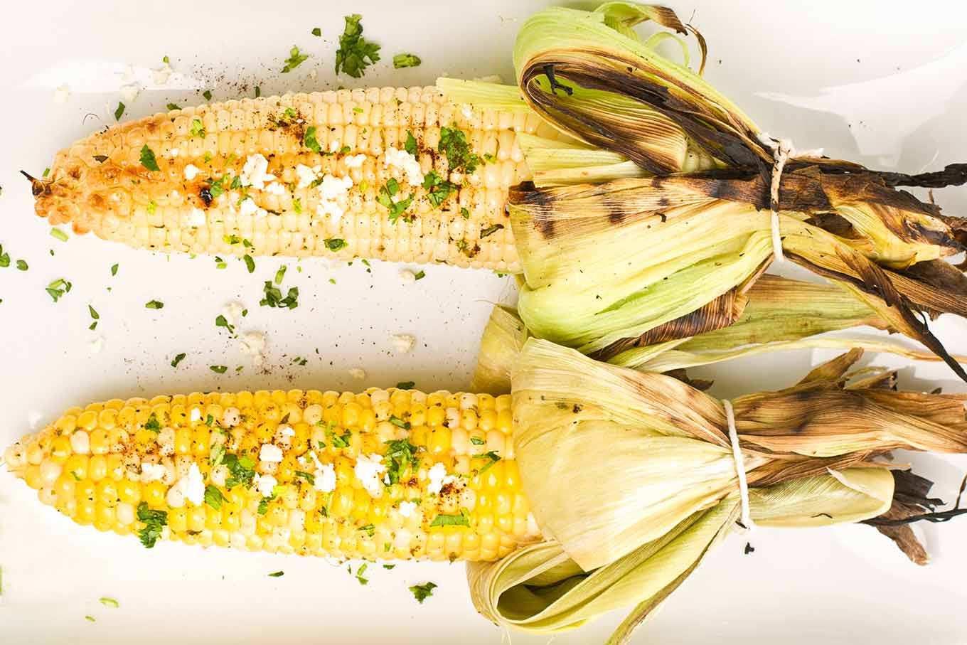 A close, overhead view of two ears of Mexican street corn ready for serving after adding melted butter, chopped cilantro, and crumbled cheese.