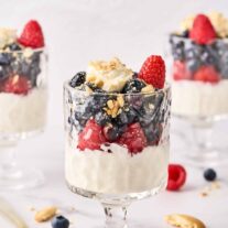Three cheesecake parfaits, each topped with blueberries, raspberries, and crumbled vanilla cookies in footed parfait glasses, surrounded by cookie crumbs, whole berries, and a spoon.